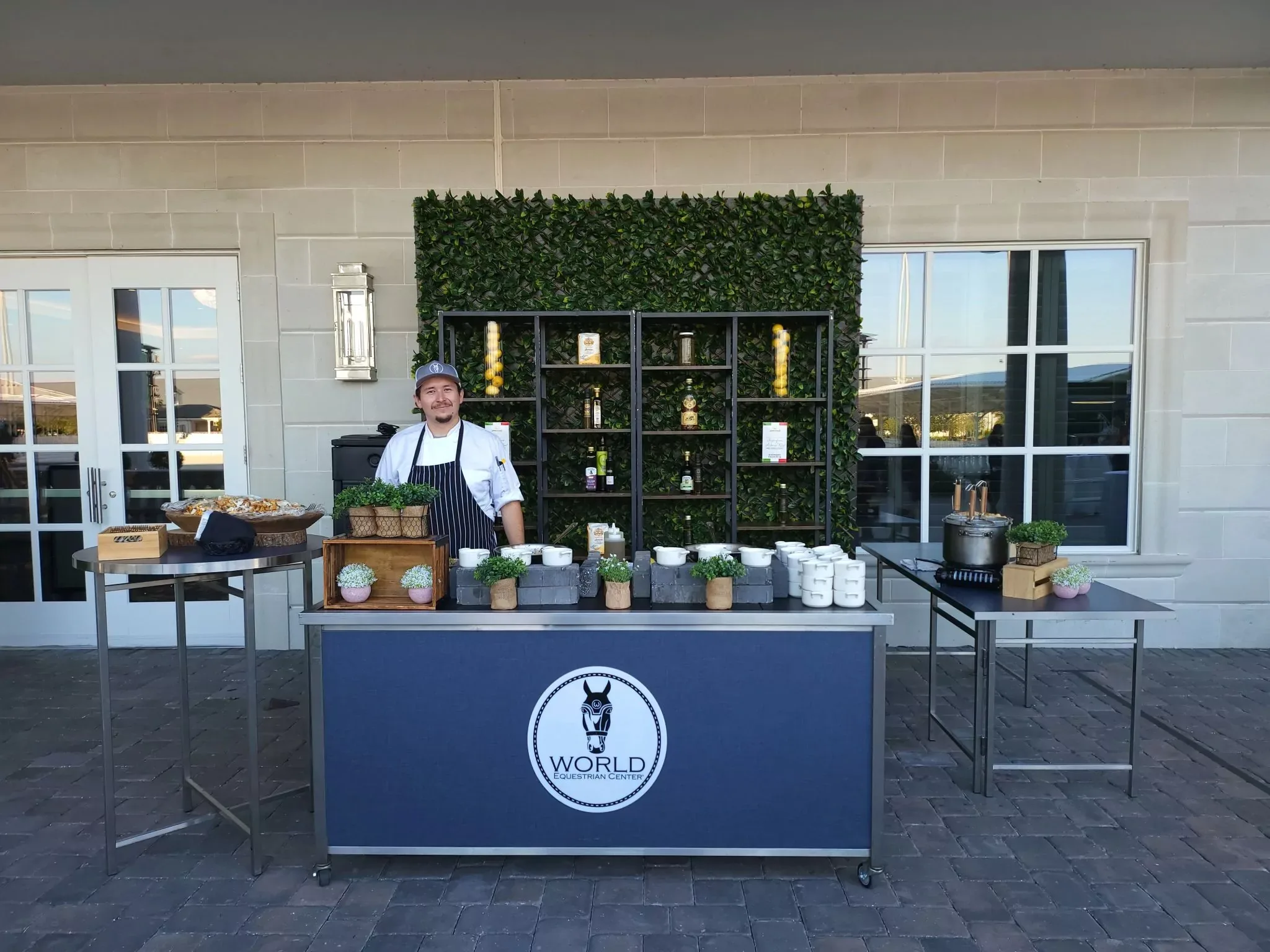A chef stands behind a table with a "World Gin" cloth, displaying herbs and drink ingredients. The setup is outdoors on a stone patio with a wall of greenery and a shelf in the background.