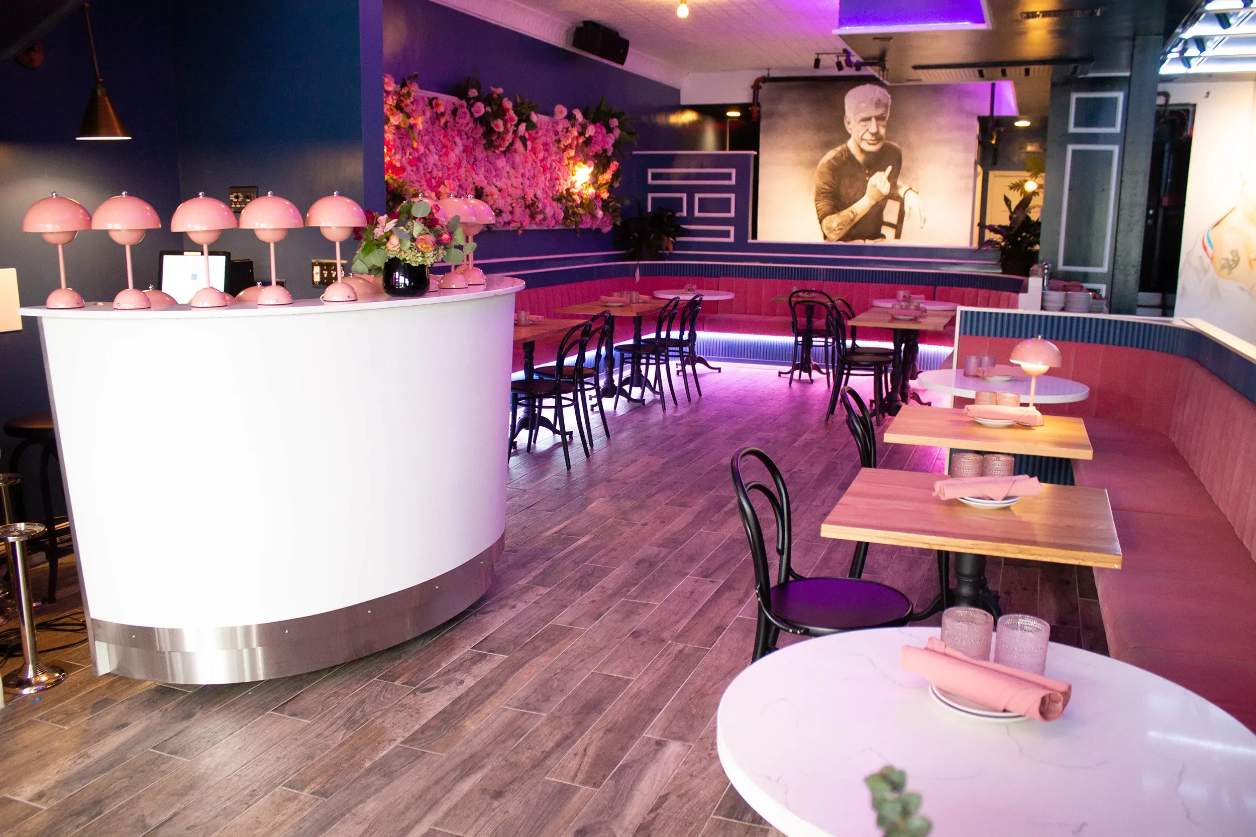 A stylish restaurant interior featuring pink and blue decor. The space has round tables with chairs, pink table lamps, and a large portrait on the wall. Bright lighting and floral arrangements add a vibrant touch to the modern design.