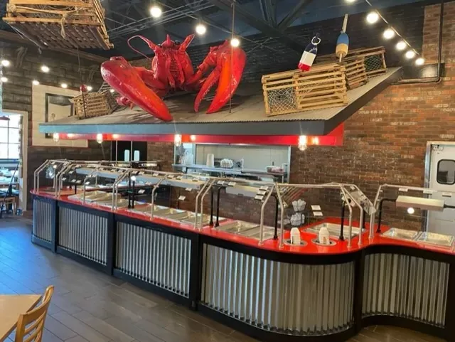 A seafood restaurant buffet with a red lobster decoration on the roof. Several empty buffet stations are lined up beneath, with nautical decor including buoys and wooden traps. Brick walls and ceiling lights create a cozy atmosphere.