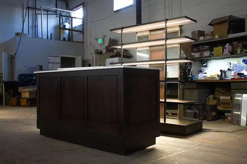 A large, dark wooden counter and illuminated shelving units in a workshop. Industrial shelves with various tools and boxes are in the background. Natural light filters through small windows.