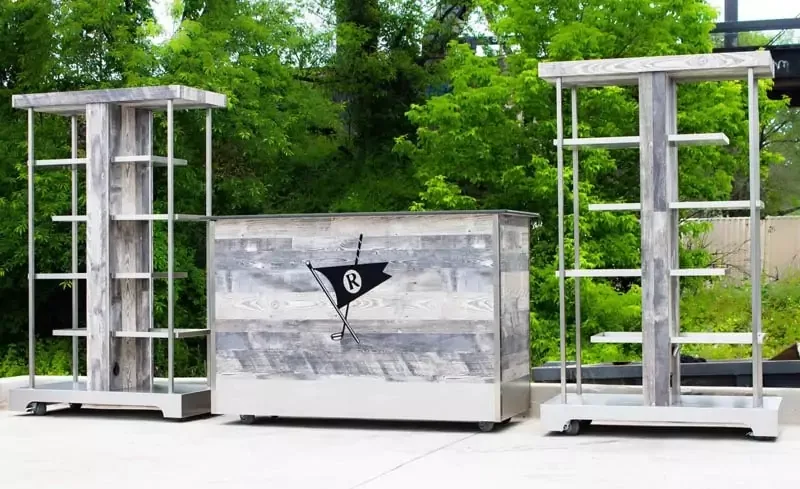 Outdoor furniture set featuring a central wooden bar with a flag logo, flanked by two tall, matching shelving units. The pieces are made of gray weathered wood and metal, set against a backdrop of lush green trees.